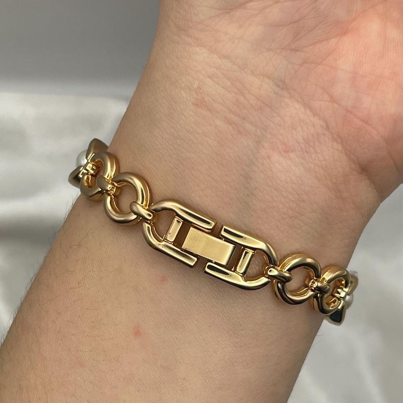 May include: Gold-toned bracelet featuring a series of interlocking links and a secure clasp. The bracelet has a polished finish and includes small, round, white pearls at the ends of some links. A classic piece of jewelry.