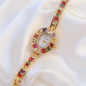 May include: A gold-tone quartz watch with an oval face featuring the text Lsotr. The bezel and bracelet are set with square-cut multicolored gemstones in shades of red, purple, yellow, and green. The watch dial has gold hands and markers.