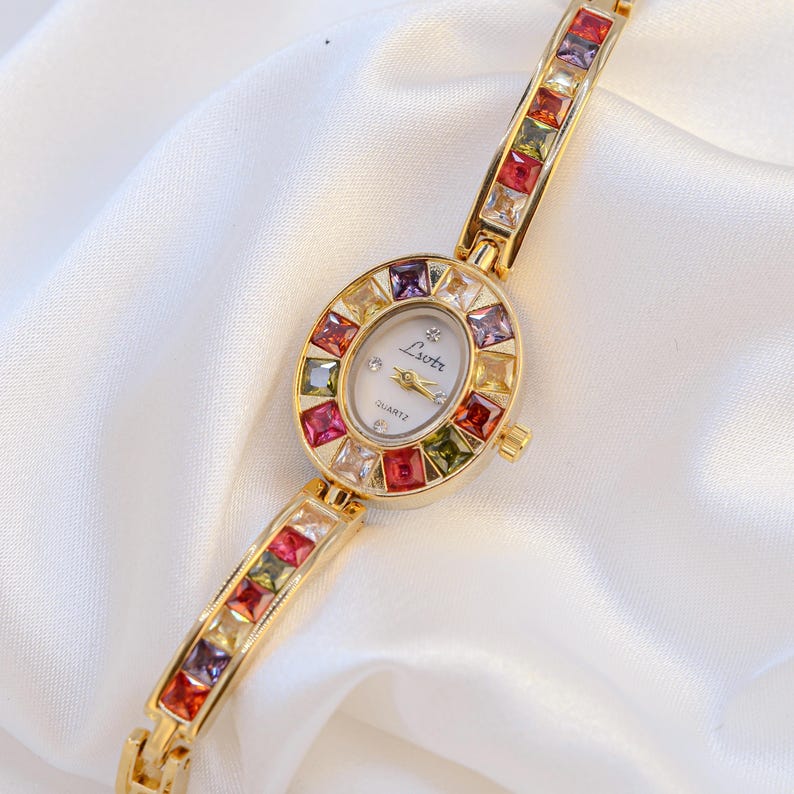 May include: A gold-tone quartz watch with an oval face featuring the text Lsotr. The bezel and bracelet are set with square-cut multicolored gemstones in shades of red, purple, yellow, and green. The watch dial has gold hands and markers.