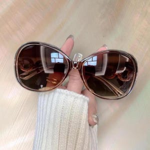 Puede incluir: Gafas de sol marrones degradadas con una montura grande y redondeada. Las lentes pasan de un tono m&aacute;s oscuro en la parte superior a un marr&oacute;n m&aacute;s claro en la parte inferior. La montura es de color marr&oacute;n claro. Las gafas de sol est&aacute;n sujetas por una mano.