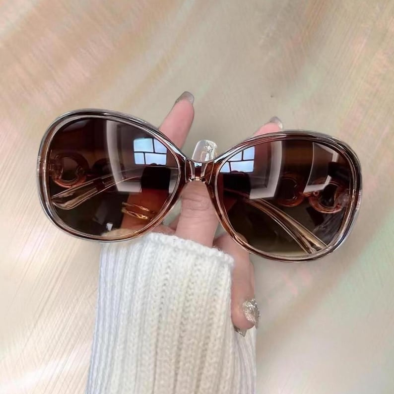 Puede incluir: Gafas de sol marrones degradadas con una montura grande y redondeada. Las lentes pasan de un tono m&aacute;s oscuro en la parte superior a un marr&oacute;n m&aacute;s claro en la parte inferior. La montura es de color marr&oacute;n claro. Las gafas de sol est&aacute;n sujetas por una mano.