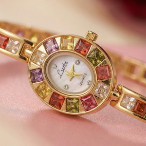 May include: An oval gold-tone quartz watch with a white mother-of-pearl dial featuring the text Lsotr. The bezel and bracelet are set with square-cut multicolored gemstones in shades of red, yellow, green, purple, and clear white.