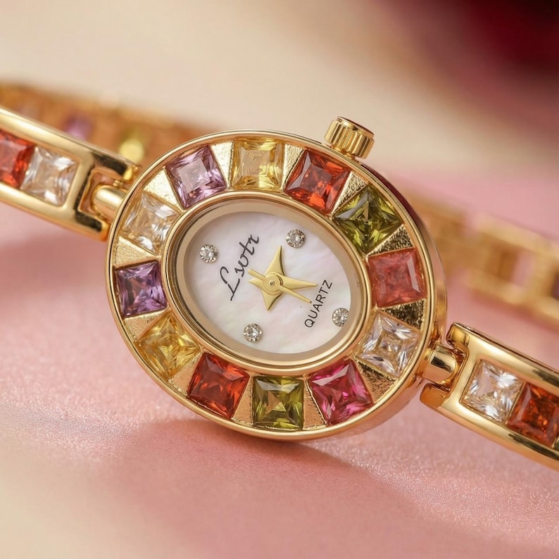 May include: An oval gold-tone quartz watch with a white mother-of-pearl dial featuring the text Lsotr. The bezel and bracelet are set with square-cut multicolored gemstones in shades of red, yellow, green, purple, and clear white.