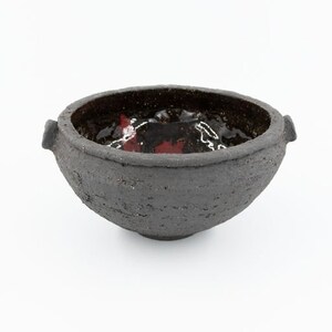 May include: A black ceramic bowl with a rough texture and a red and black glaze inside. The bowl has two small handles on the sides.