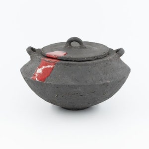 May include: A black ceramic pot with a lid, featuring a red glaze detail. The pot has a rounded shape and a small handle on the lid.
