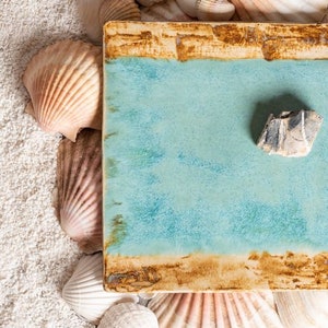 Könnte beinhalten: Ein blaues Keramiktablett mit rustikalem braunem Rand liegt auf einem Bett aus weißem Sand. Das Tablett ist von Muscheln und einem kleinen grauen Stein umgeben.
