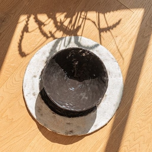 May include: A black ceramic bowl with a white rim sits on a white ceramic plate with a speckled glaze. The bowl and plate are on a wooden surface.