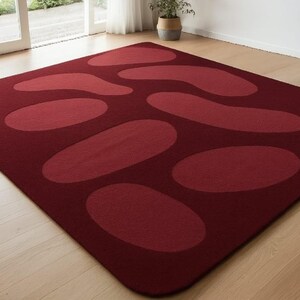 May include: A rectangular burgundy rug with a pattern of large, lighter red oval and amoeba-shaped designs. The rug is placed on a light-colored wooden floor, with a window and a potted plant visible in the background.