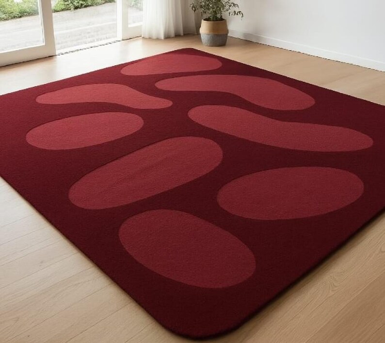 May include: A rectangular burgundy rug with a pattern of large, lighter red oval and amoeba-shaped designs. The rug is placed on a light-colored wooden floor, with a window and a potted plant visible in the background.