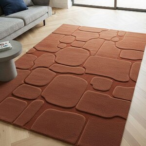 May include: A rectangular, rust-colored rug with a raised stone pattern. The rug features various sizes and shapes of stones, creating a textured, three-dimensional effect. The rug is placed on a light-colored wooden floor, with a gray sofa and side table in the background.