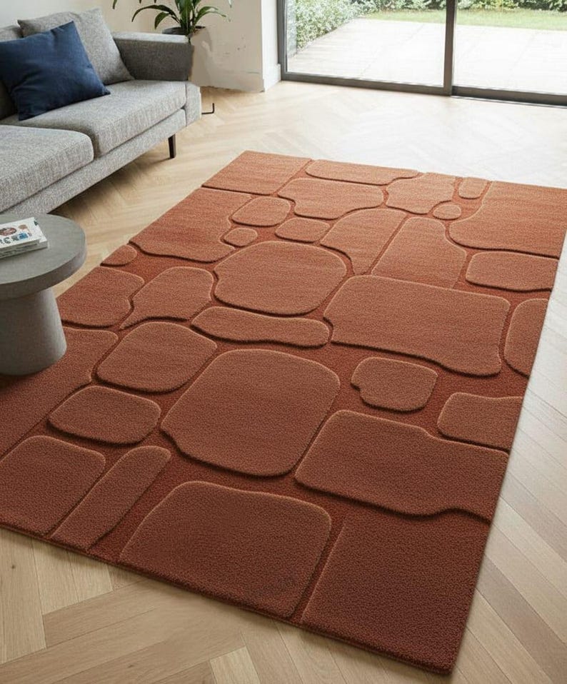 May include: A rectangular, rust-colored rug with a raised stone pattern. The rug features various sizes and shapes of stones, creating a textured, three-dimensional effect. The rug is placed on a light-colored wooden floor, with a gray sofa and side table in the background.