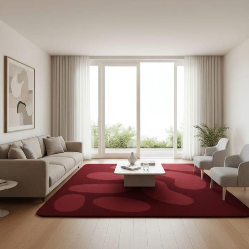May include: A modern living room featuring a large burgundy rug with an abstract pattern. The room includes a beige sofa, two gray armchairs, a white coffee table, and a large window with sheer white curtains. A framed artwork hangs on the wall.