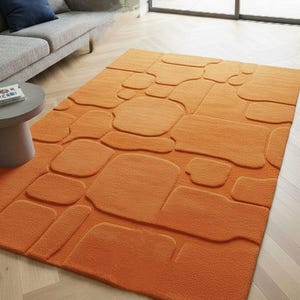 May include: An orange rectangular rug with a raised, stone-like pattern. The rug's design features various shapes and sizes, creating a textured appearance. The rug is placed on a light-colored wooden floor, and is near a gray sofa.