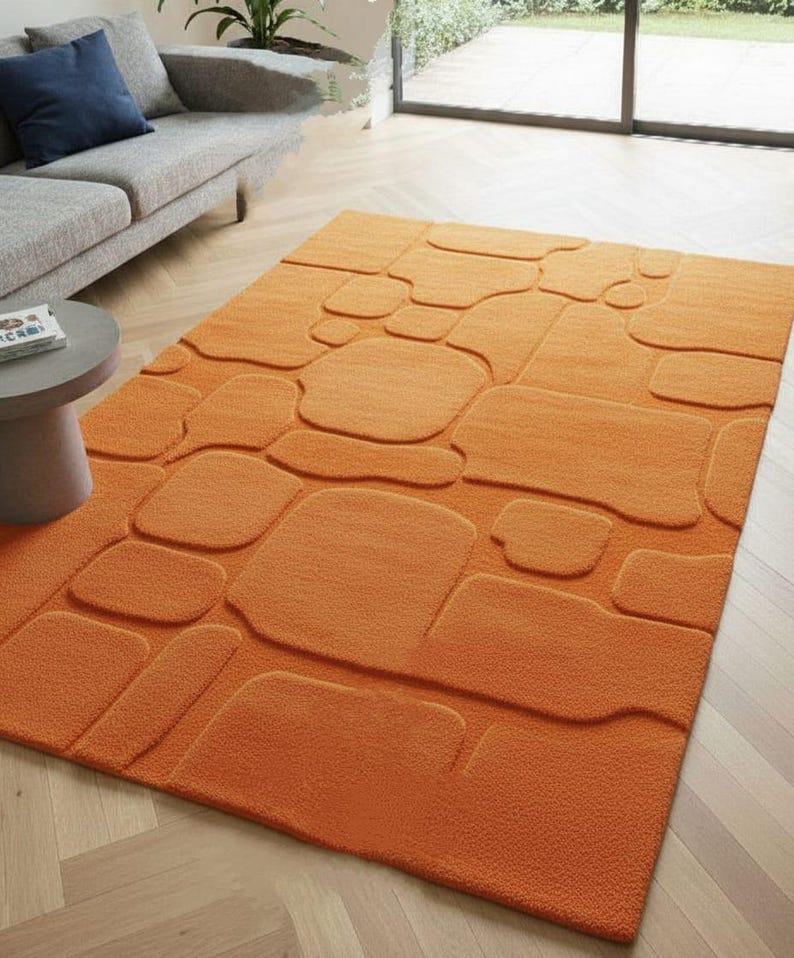 May include: An orange rectangular rug with a raised, stone-like pattern. The rug's design features various shapes and sizes, creating a textured appearance. The rug is placed on a light-colored wooden floor, and is near a gray sofa.