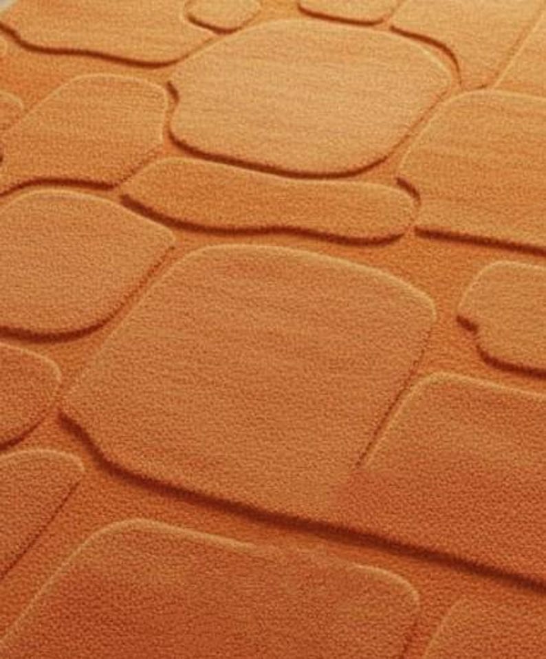 May include: Close-up of an orange bath mat with a stone-like pattern. The mat features raised, rounded shapes resembling cobblestones, creating a textured surface. The color is a warm, earthy orange.