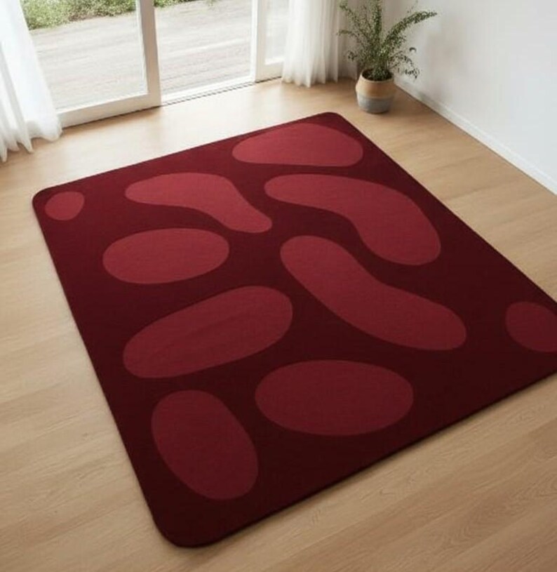 May include: A rectangular burgundy rug with a pattern of large, lighter burgundy organic shapes. The rug is placed on a light-colored wooden floor, with a window and a potted plant in the background.