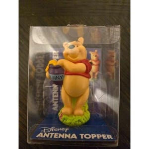 May include: A Winnie the Pooh antenna topper figurine in a clear plastic case. The yellow bear is wearing a red shirt and holding a purple honey pot. The base is green and blue, with the text "Disney Antenna Topper".