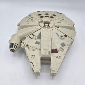 Könnte beinhalten: Ein Kunststoff-Spielzeugmodell des Millennium Falcon Raumschiffs mit einem kreisförmigen, cremefarbenen Rumpf, geformten mechanischen Details, einem seitlich angebrachten Cockpit mit schwarzen Fenstern sowie kleinen kastanienbraunen und grauen Akzenten.