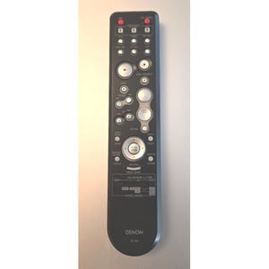 Op de afbeelding: Een zwarte Denon RC-1098 afstandsbediening met talloze zilveren knoppen, twee rode aan-uitknoppen aan de bovenkant en een centrale navigatieknop. Het apparaat bevat diverse gelabelde bedieningselementen voor bronselectie en volumeregeling.