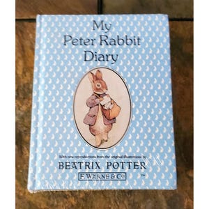 Op de afbeelding: Een hardcover "My Peter Rabbit Diary" boek. De omslag is lichtblauw met een herhalend patroon van witte konijn silhouetten. Het boek bevat een illustratie van Peter Rabbit en tekst van Beatrix Potter.