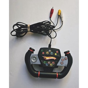 May include: Black Hot Wheels steering wheel controller with red and silver accents. Features multiple colored buttons and the Hot Wheels logo. Includes a black cable with red, yellow, and white RCA connectors.