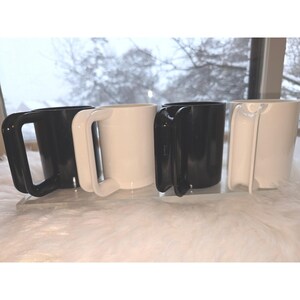 Puede incluir: Cuatro tazas de cerámica, dos negras y dos blancas, se exhiben sobre un soporte de acrílico transparente. Cada taza tiene un diseño de asa único. Las tazas están sobre un fondo borroso de invierno.