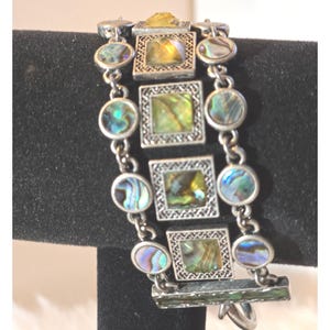 Op de afbeelding: Zilverkleurige armband met vierkante en ovale abalone schelp accenten. De armband heeft een rechthoekige sluiting en is samengesteld uit verbonden secties met decoratieve frames rond de abalone schelp stukken. De abalone schelpen tonen iriserende kleuren van groen, blauw en goud.