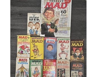 May include: A collection of vintage "MAD" magazine books, featuring various covers with the iconic Alfred E. Neuman. The books showcase vibrant colors and bold typography, with titles like "Totally MAD" and "Fighting MAD."