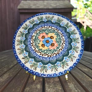 May include: A round ceramic plate featuring a central cluster of four painted flowers in shades of peach, yellow, and blue. The design includes intricate green foliage, brown swirls, and a decorative blue and white border pattern around the edge.