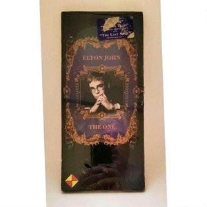 May include: A black CD case featuring Elton John's album "The One." The cover has a portrait of Elton John in glasses, framed by ornate gold and purple designs. The album title is displayed below the portrait.