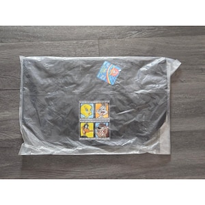 May include: A black item, possibly a bag or blanket, sealed in clear plastic. A colorful patch featuring Looney Tunes characters like Tweety Bird, Bugs Bunny, Daffy Duck, and Taz is visible. A small tag is attached to the plastic.