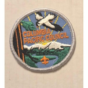 May include: A round patch with a blue background and a silver border. The patch features a flying bird, mountains, and the text "COLUMBIA PACIFIC COUNCIL" in orange. A green tree and a fleur-de-lis are also present.