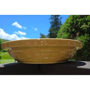 Może przedstawiać: Okrągła, musztardowo-żółta ceramiczna forma do pieczenia ciasta z błyszczącym wykończeniem. Zewnętrzna strona posiada dekoracyjny, tłoczony wzór plecionki na bokach, a górna krawędź ma falisty, żłobkowany design dla klasycznego i funkcjonalnego wyglądu.