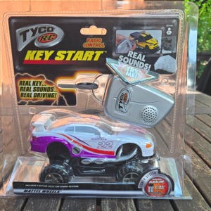 May include: A Tyco RC Key Start radio-controlled toy car in its original plastic packaging. The white and purple Mustang features the number 22, a rear spoiler, and large black tires. Includes a silver key-shaped remote control transmitter.