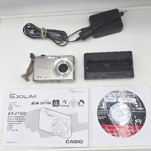 May include: A silver Casio Exilim EX-Z1000 digital camera with a wrist strap, a black charging dock, a black power adapter with a cord, a printed basic reference manual, and a software disc. The camera features 10.1 megapixels printed on the front.