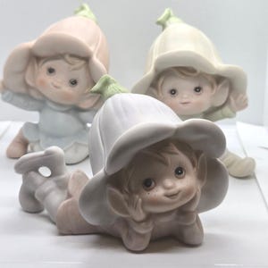 May include: Three porcelain figurines of small elf-like creatures with pointed ears and freckled faces. Each figure wears a large, bell-shaped flower hat in shades of pale pink, cream, or white, with a small green stem detail on top.