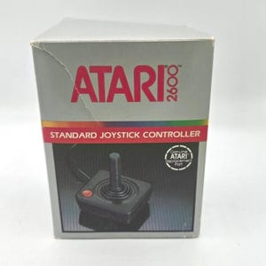 May include: A grey Atari 2600 box with a red Atari logo. The box displays a photo of a black joystick controller with a red button. Text on the box reads "STANDARD JOYSTICK CONTROLLER" and "Genuine Atari Replacement Part."