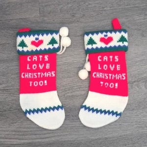 May include: Two red and white knit Christmas stockings featuring the text "CATS LOVE CHRISTMAS TOO!" in white letters. The cuffs are white with green pine trees, red hearts, and blue zigzag patterns. Each stocking includes white pom-pom accents.