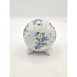 May include: A white ceramic sphere with three small feet and several small holes. The sphere is decorated with blue floral patterns. The design features clusters of blue flowers and green leaves. The object is likely a decorative item or a small container.