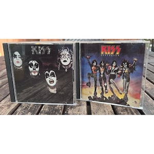 May include: Two music CDs in clear plastic cases. The left case features the band KISS with four members in black and white makeup. The right case is the Destroyer album, showing the four band members in stage costumes against a dark sky.