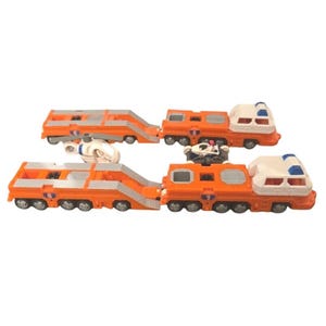 May include: Two orange and white toy transport trucks with flatbed trailers, each featuring blue accents and the number one printed on the side. A small white toy helicopter and a black and white toy race car are positioned between the two trucks.