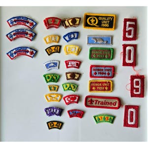 Könnte beinhalten: Eine Sammlung gestickter Aufnäher in verschiedenen Formen und Farben. Einige Aufnäher tragen Texte wie "Helping Seeing", "Quality Unit 1986", "Honor Unit 1983/1984/1982" und "Trained". Die Zahlen 5, 0, 9 und 0 sind ebenfalls vorhanden.