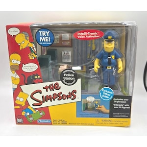 May include: The Simpsons interactive police station environment toy set. The packaging features cartoon characters and the text "The Simpsons". Includes an Officer Eddie figure and the text "Try Me!" on the side panel.