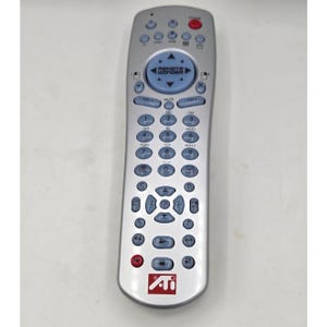 Puede incluir: Un mando a distancia de televisión plateado Remote Wonder con botones azul claro, un panel direccional circular con flechas, un botón de encendido rojo y un logotipo ATI rojo en la parte inferior. El dispositivo incluye un teclado numérico y controles multimedia.