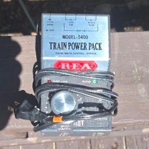 May include: A silver and gray Train Power Pack, model 5400, with the text "TRAIN POWER PACK" and "REA" printed on the front. The power pack has a dial for speed control and is wrapped with a black power cord. The power pack is on a wooden surface.