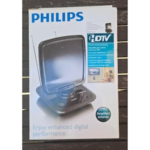 May include: A Philips indoor HDTV antenna in a white retail box. The black antenna features a square panel, two thin metal telescopic rods, and a base with a power button. The packaging displays the text Philips, HDTV, and Amplified antenna.