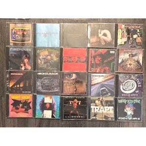 Op de afbeelding: Een verzameling van twintig muziek-cd's in plastic doosjes, gerangschikt in een raster van vier bij vijf. De hoezen bevatten diverse grafische ontwerpen, bandfoto's en tekst, waaronder titels als Nickelback, Creed, Kid Rock en Sammy Hagar.