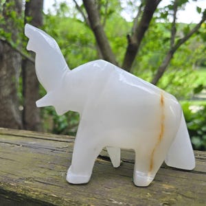 May include: A white, carved stone elephant figurine with its trunk raised. The sculpture has a smooth, polished finish and a subtle, natural brown streak down its side. The elephant is standing on a weathered wooden surface, with a blurred green background.