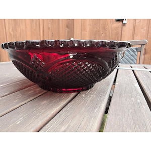 Puede incluir: Un gran cuenco de vidrio rojo rubí con un borde festoneado. El cuenco presenta un patrón de corte de diamante y un diseño decorativo en los laterales. Se muestra sobre una mesa de madera, mostrando su rico color y detalles intrincados.