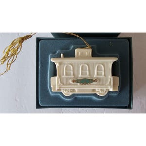 May include: Ivory train caboose ornament with three arched windows and a green and gold decorative accent. The ornament is displayed in a teal box with a gold tassel attached.